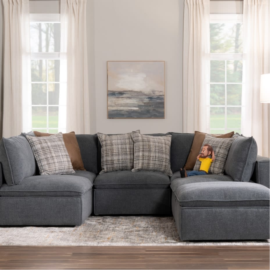 Modern living room with a large gray fabric sectional sofa featuring multiple patterned and solid throw pillows, a neutral-toned abstract wall art, and natural light from tall windows.