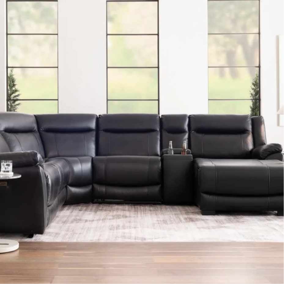 Black leather sectional sofa with reclining seats, cup holders, and modern stitching, set in a bright living room with large windows.