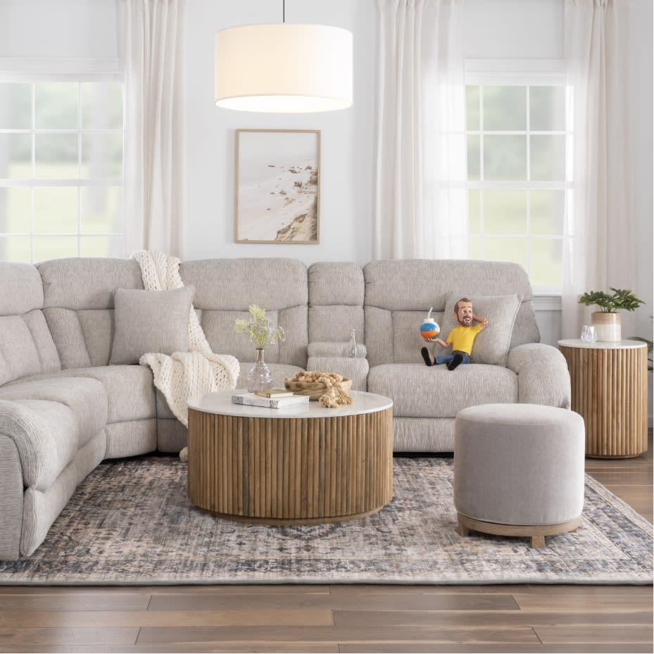Light gray sectional sofa with plush cushions and a knitted throw, paired with round wooden coffee and side tables, a gray ottoman, and large windows with white curtains in a bright living room.