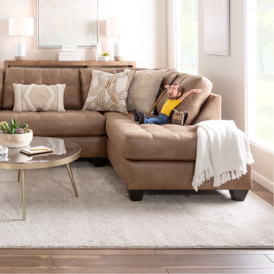 Brown leather sectional sofa with decorative beige and patterned pillows, white throw blanket, glass coffee table, and wooden floor in a bright living room.