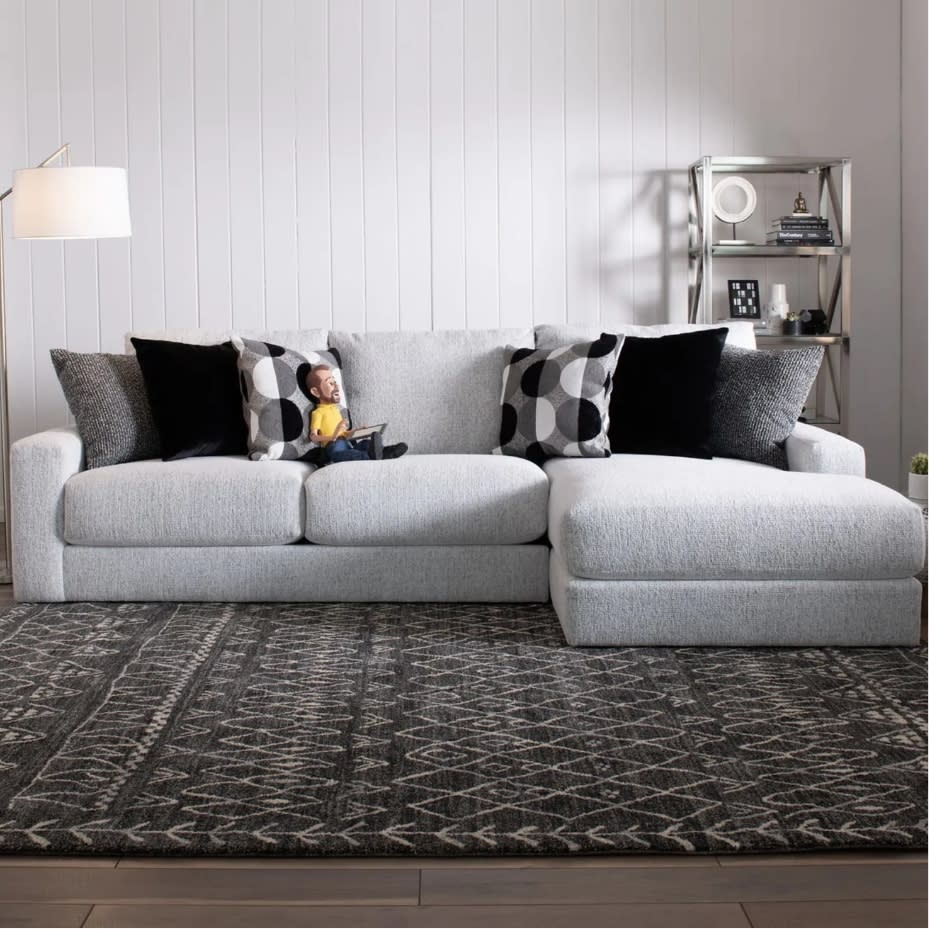 Light gray fabric sectional sofa with chaise, featuring black and patterned throw pillows, a patterned dark rug, modern floor lamp, and metal shelving unit.