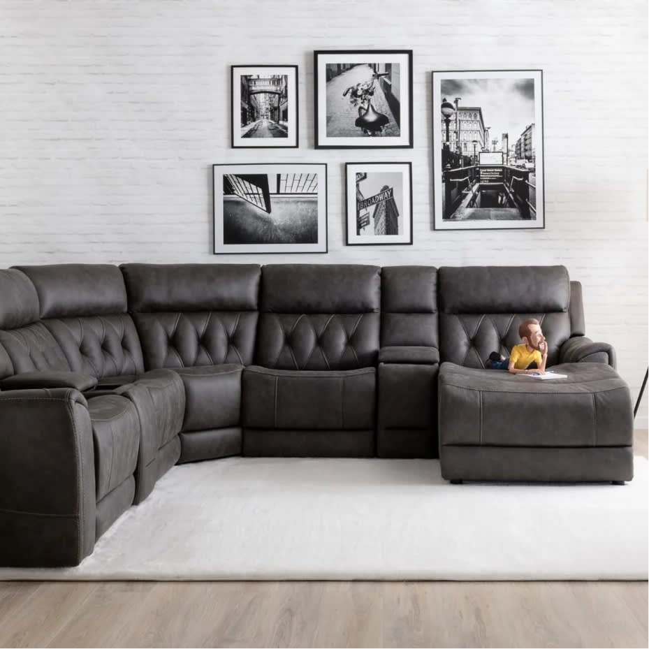 Dark gray leather sectional sofa with tufted cushions and reclining seats, set against a white brick wall with black-and-white framed cityscape photos.