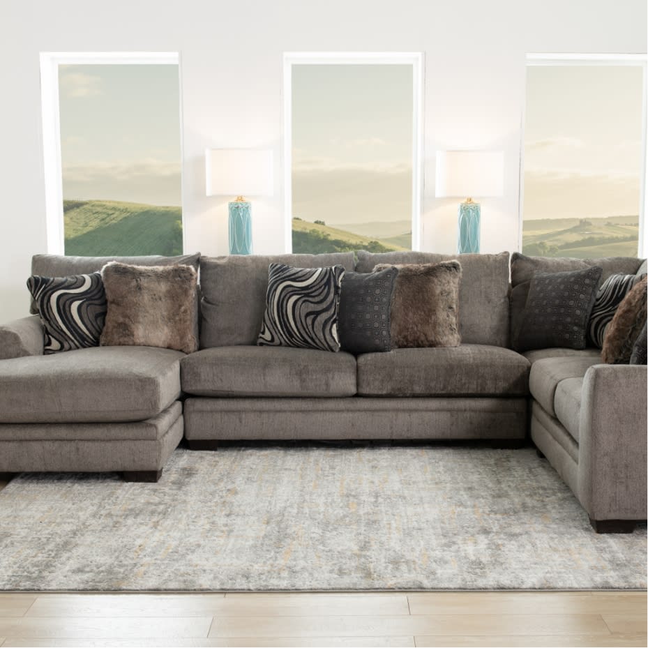 Gray fabric sectional sofa with plush cushions and patterned throw pillows, positioned in a bright living room with three tall windows and blue ceramic lamps.