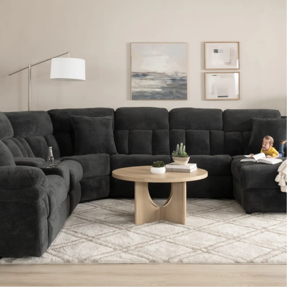 Dark gray sectional sofa with plush cushions and reclining seats, centered around a round light wood coffee table on a patterned beige rug, with modern abstract wall art and a floor lamp in a neutral-toned living room.