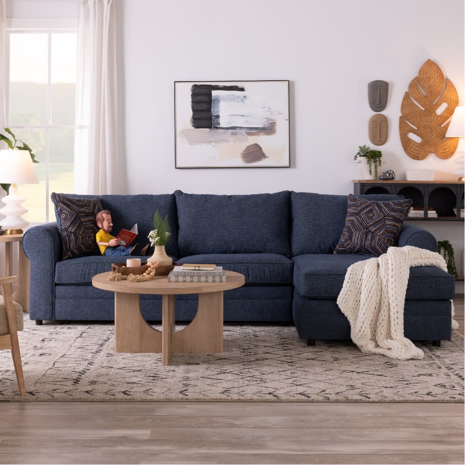 Dark blue fabric sectional sofa with chaise, patterned throw pillows, cream knit blanket, wooden round coffee table with books and decor, modern living room setting.