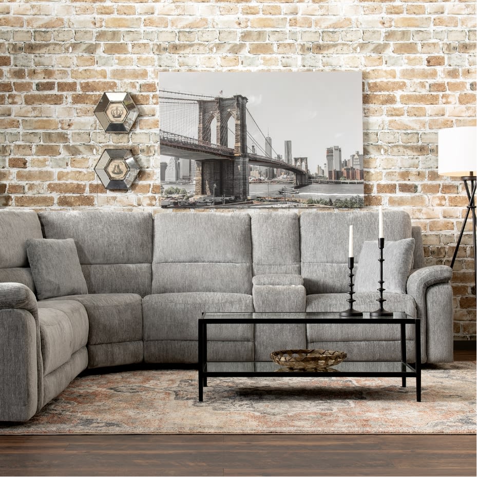 Modern living room with a gray fabric sectional sofa, black metal and glass coffee table, beige patterned rug, exposed brick wall, and a black-and-white Brooklyn Bridge wall art.