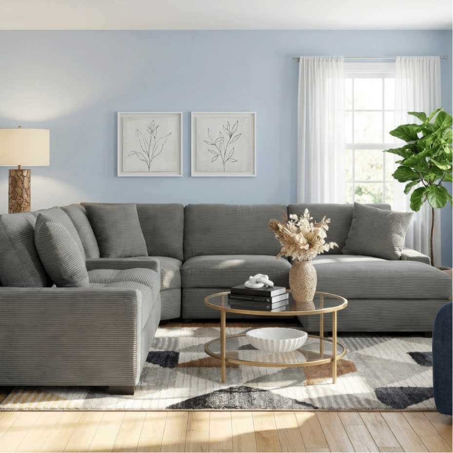 Modern living room with a gray corduroy sectional sofa, round glass coffee table with gold frame, neutral abstract rug, and natural light from window.