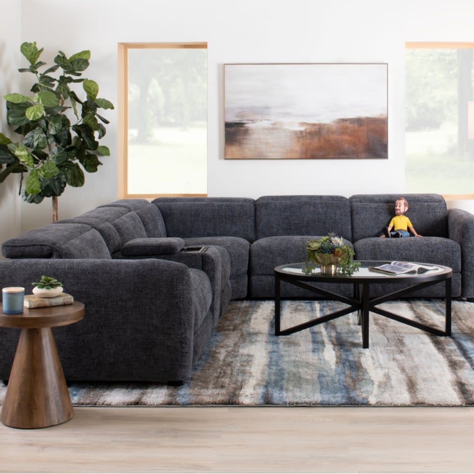 Dark gray sectional sofa with plush fabric upholstery in a modern living room, featuring a round black metal coffee table, wooden side table, abstract wall art, and large green plant.