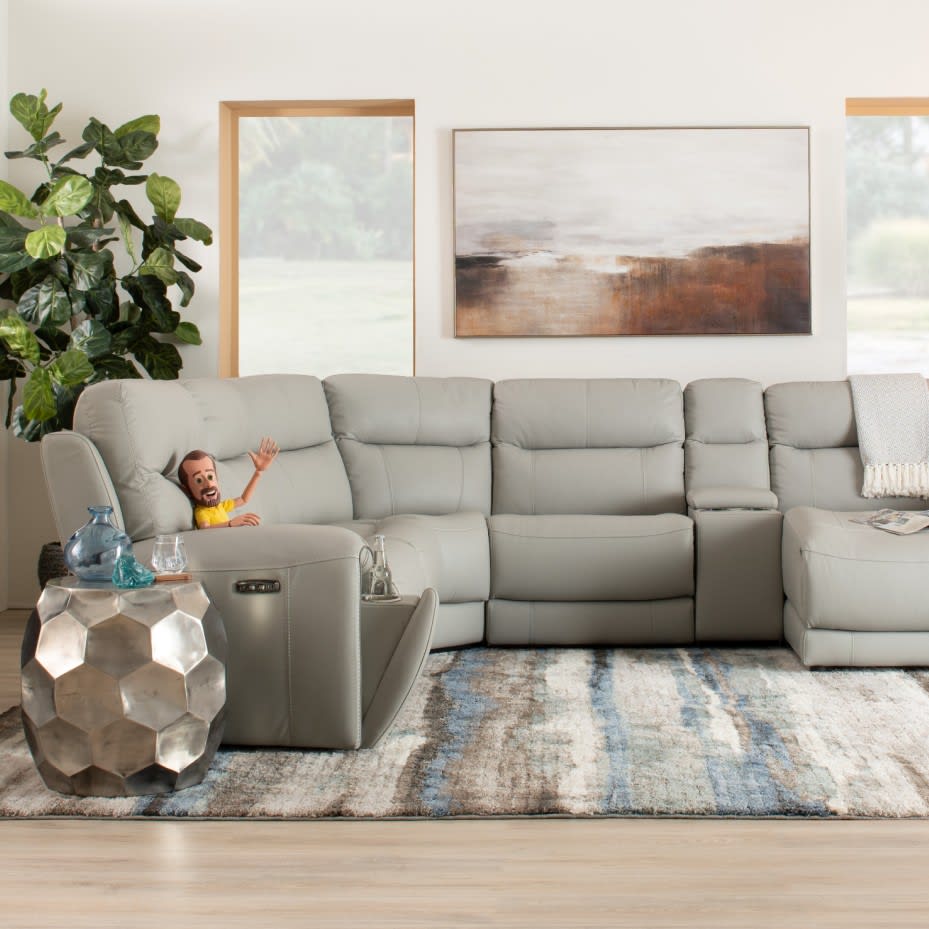Modern light gray leather sectional sofa with recliner and cup holder, set on a blue and beige patterned rug in a bright living room with large windows and abstract wall art.