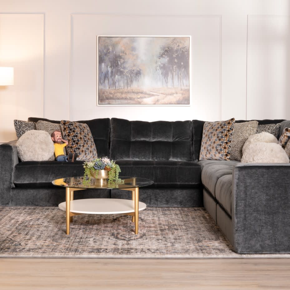 Dark gray velvet sectional sofa with tufted cushions, patterned and fluffy throw pillows, a round glass coffee table with gold legs, and a muted area rug in a modern living room.
