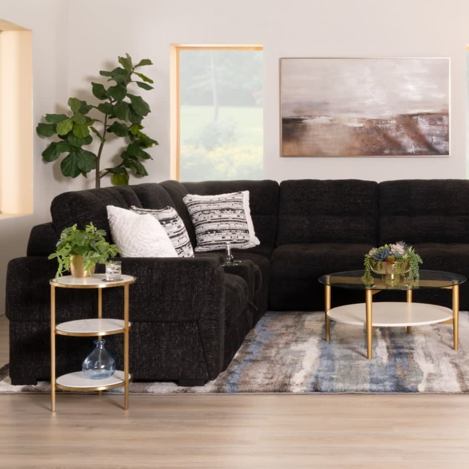 Black sectional sofa with textured fabric, patterned throw pillows, gold metal side and coffee tables with glass tops, decorative plants, and abstract wall art in a modern living room.