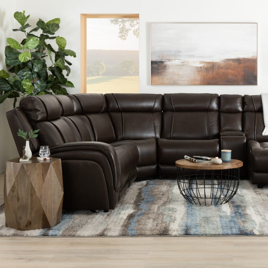 Dark brown leather sectional sofa with plush cushioning and reclining seats in a modern living room, paired with a geometric wooden side table, round wireframe coffee table with wooden top, abstract wall art, and a large green plant.