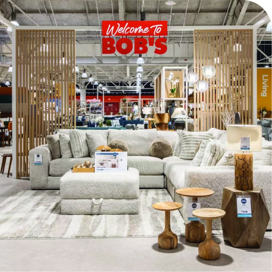 Image of a Bob's Discount Furniture store with a light gray sectional sofa round wood accent tables and a gray and a white striped rug.