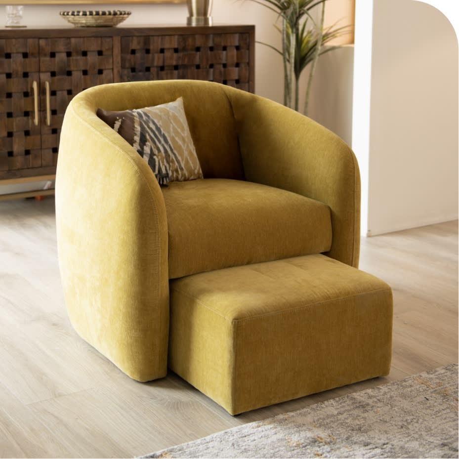 Modern mustard yellow velvet armchair with matching ottoman, curved design, and decorative pillows, set on light wood flooring.