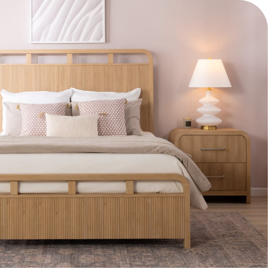 Queen-sized bed with light wood slatted headboard and footboard, beige bedding, decorative pillows, matching wood nightstand with white lamp, and neutral-toned rug.