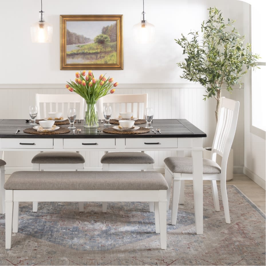 Dining room setup with a black wooden table, white cushioned chairs and bench, gray woven seat cushions, tulip centerpiece, pendant lights, and framed landscape art.