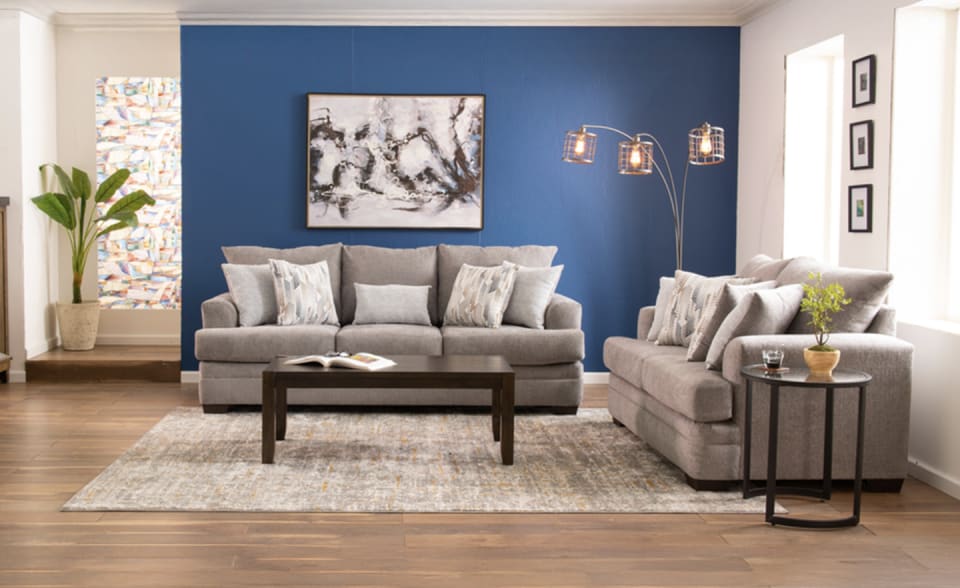 A cozy living room features a gray sofa set with patterned pillows, a dark wood coffee table, and a modern floor lamp against a blue accent wall and abstract art.