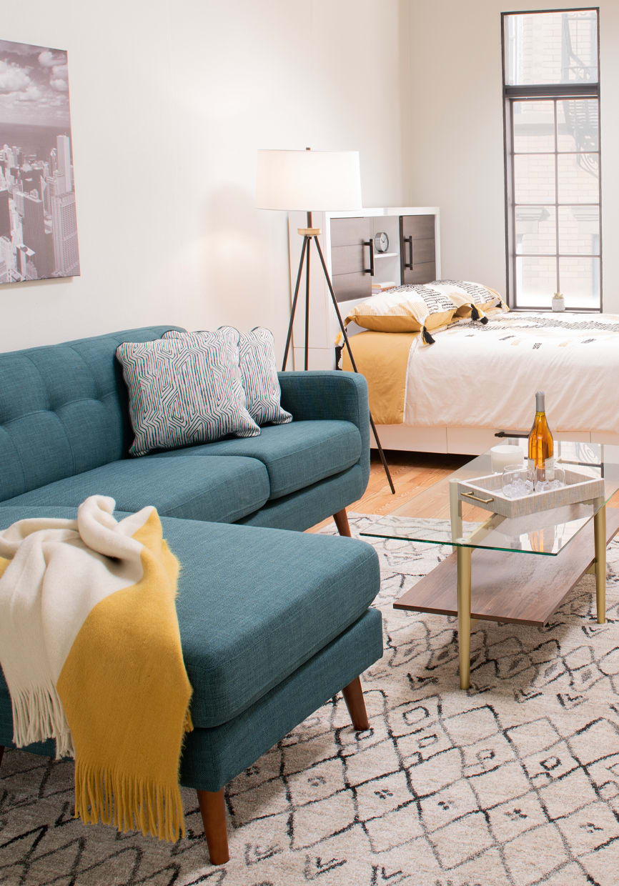 Small space living space with blue chofa with accent pillows, and throw blanket. Beige and gray area rug with glass top coffee table and white bed in the background.
