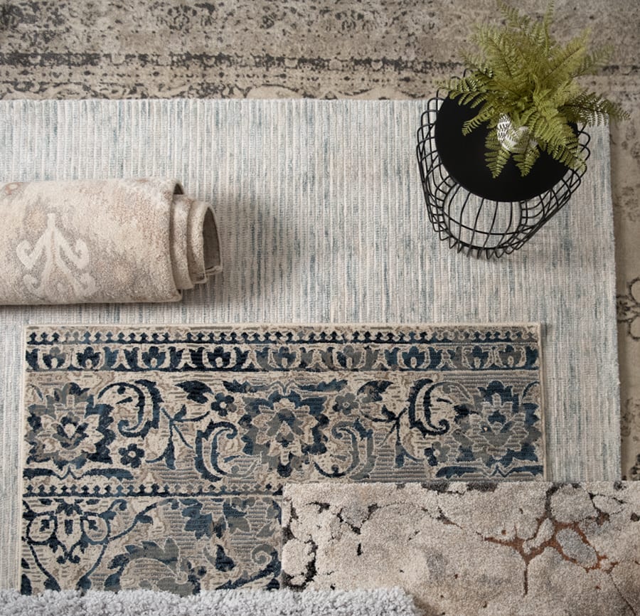 5 rugs overlapping one another that have tones of blue and beige. Each rug has a different pattern. A plant is on the larger area rug.