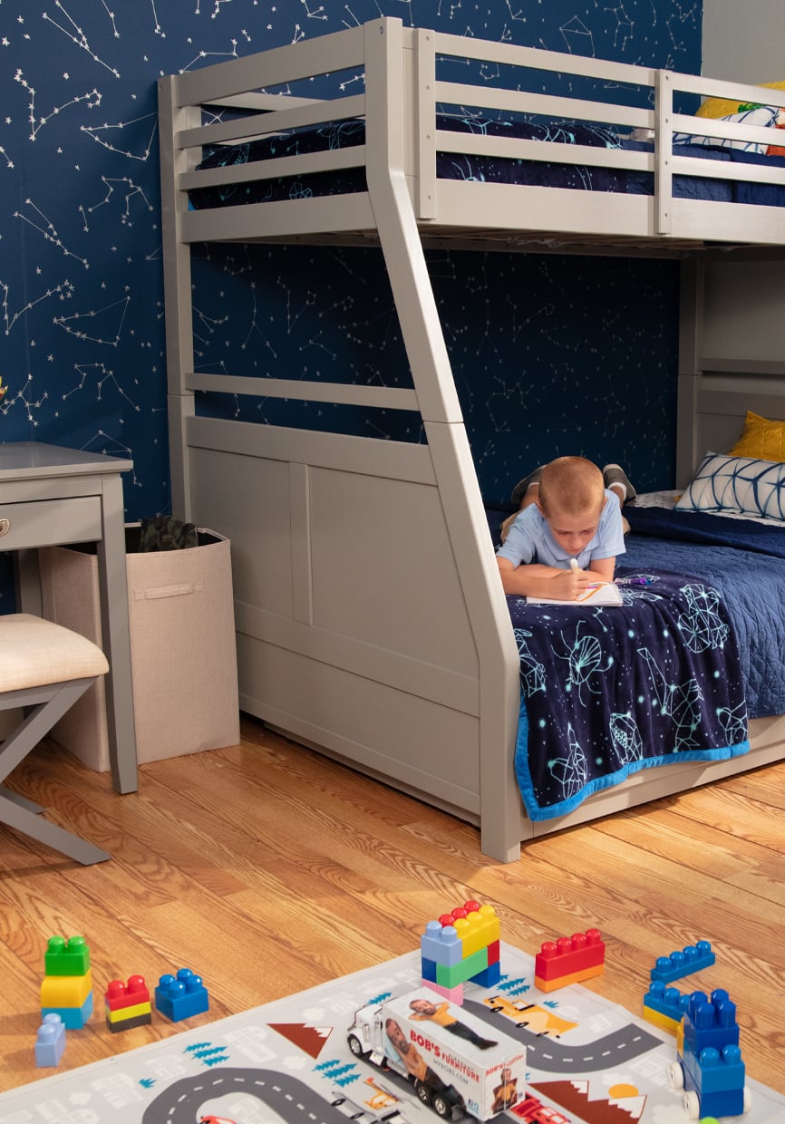 Gray twin over full size bunkbed with child relaxing in the bottom bunk. Gray desk and chair, hammper, play rug, and toys decorate the room.