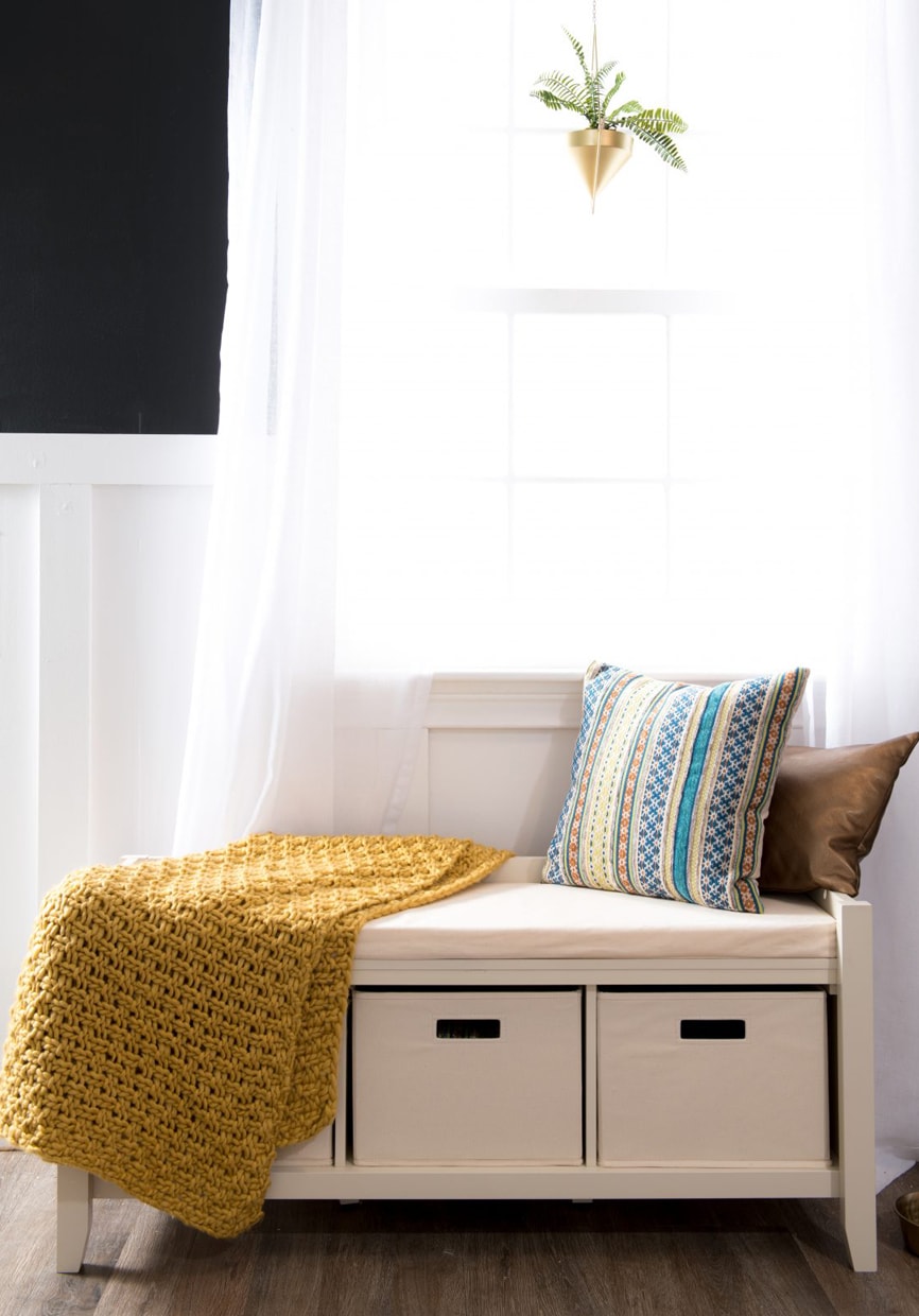 White storage bench with mustard yellow throw blanket, brown lumbar pillow, and blue, gray, and white striped accent pillow.