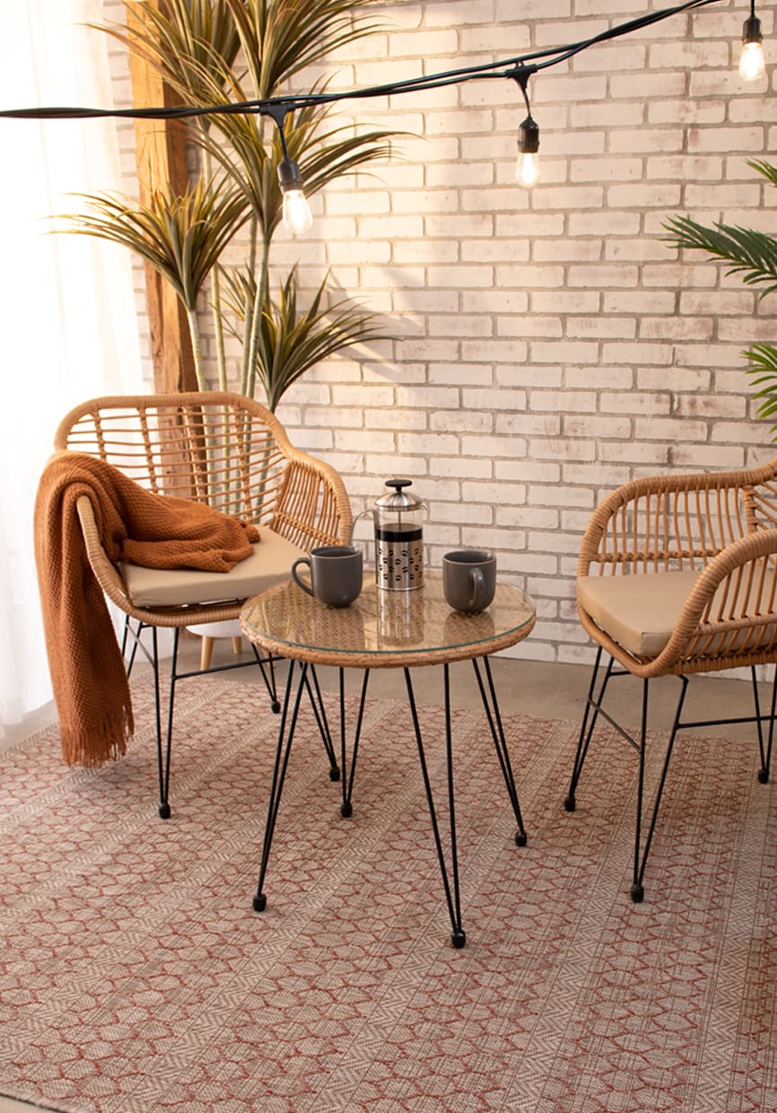 2 brown wicker outdoor chairs with white cushions, and orange throw blanket, and a brown wicker glass top bistro table centered on an abstract ivory and burnt orange abstract rug with outdoor string lights overhead.