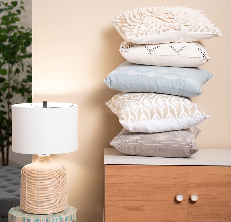 Beige table lamp on round accent table that has a mix of pastel colors. Wooden accent storage cabinet with white top and 5 different accent pillows stacked on top.