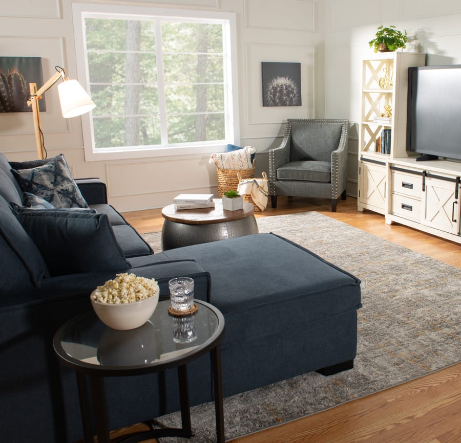 Small space living room with a blue 2 piece left arm facing chaise sectional and a glass side table. Round silver storage ottoman with wooden top. White media center with storage shelves and cabinets.