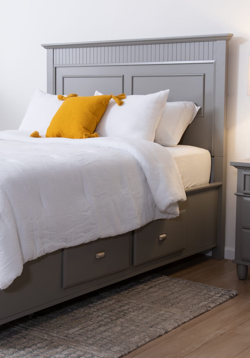 Gray panel storage bed with white bedding and a mustard yellow accent pillow. Gray plush rug anchors the bedroom.