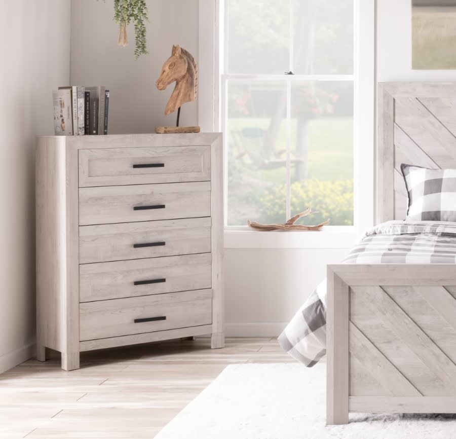 White bedroom chest with 5 drawers in the corner of a bedroom and matching bed with plaid bedding.