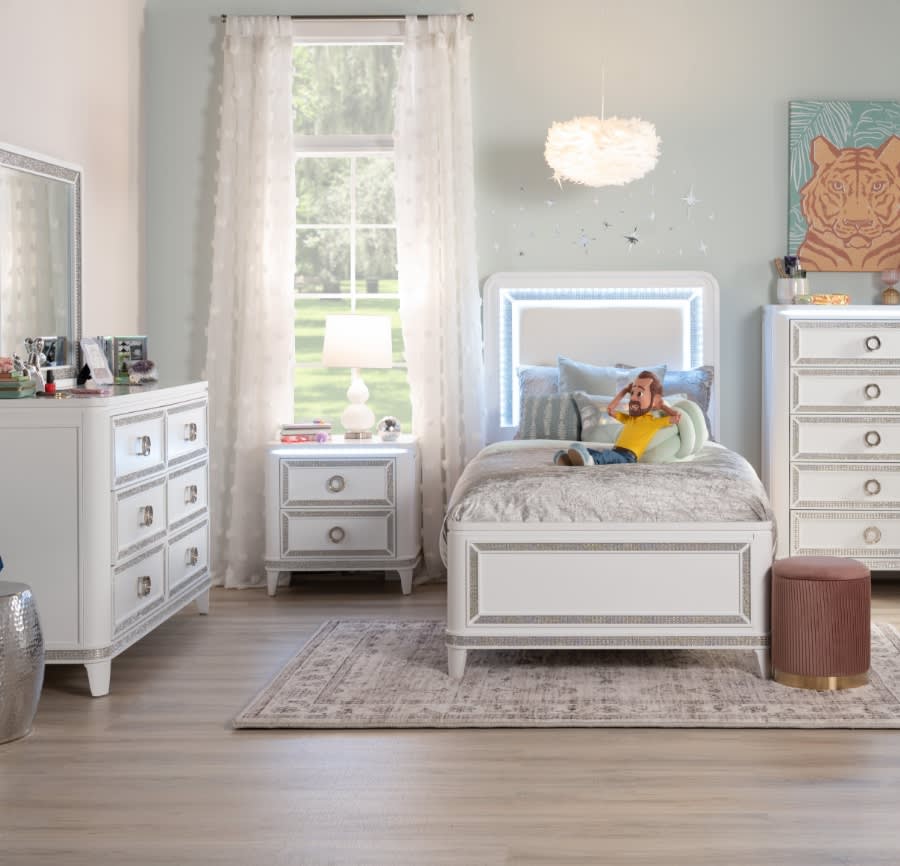 White twin sized bed with illuminated headboard, 6 drawer dresser with mirror, 5 drawer chest, and 2 drawer nightstand on a gray area rug. A fluffy pendant light, abstract tiger artwork, and blush upholstered ottoman decorate the space.