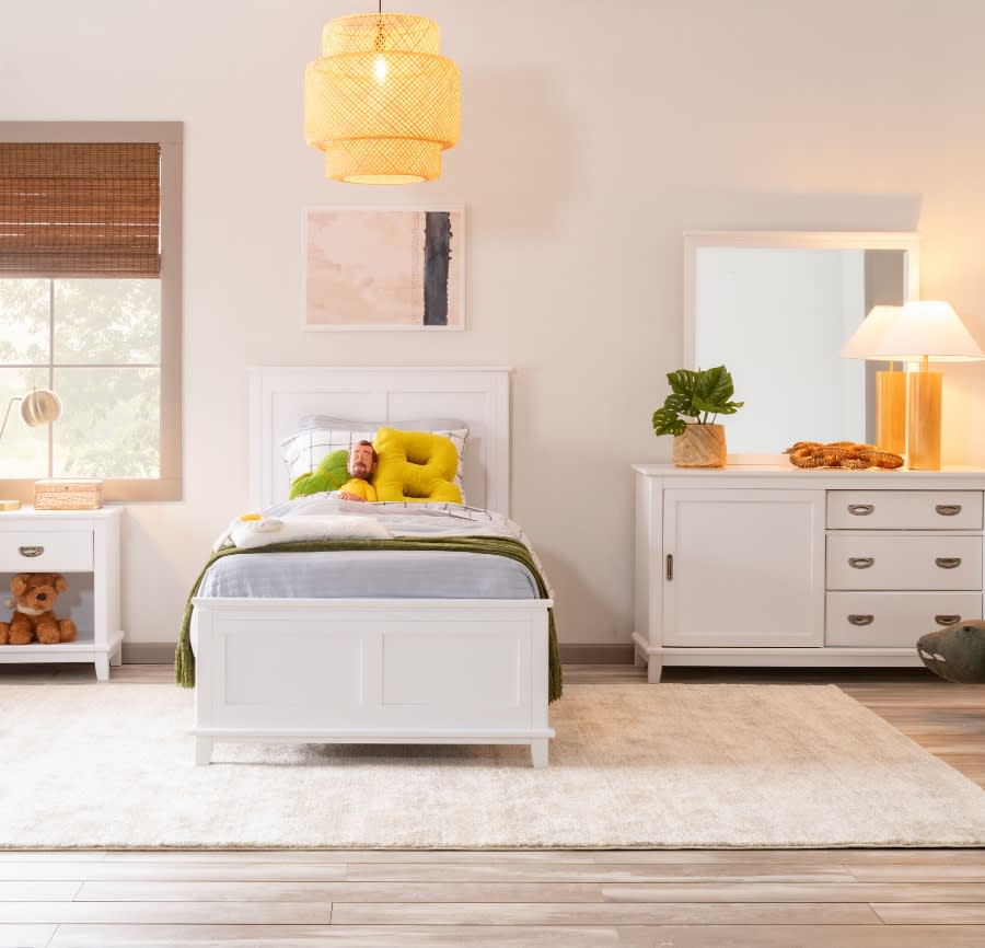 Kids bedroom with white twin sized bed, 3 drawer dresser with mirror and cabinet storage, white nightstand with drawer positioned on a white patterned rug.