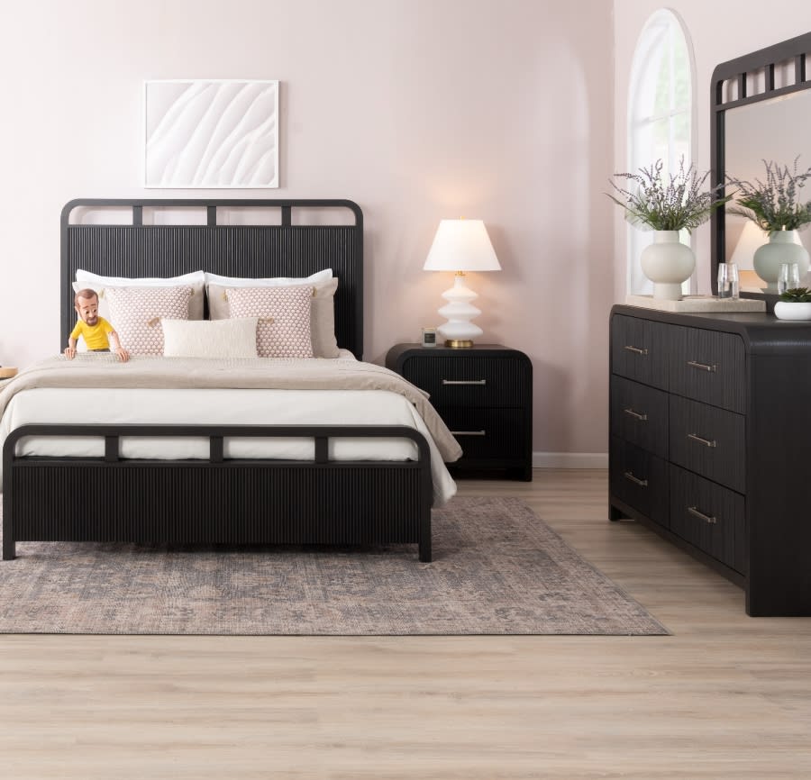 Transitional bedroom set in black featuring a bed with headboard with white sheets and beige comforter, 6 drawer dresser and mirror, and 2 drawer nightstand with white table lamp, all against a white wall and on top a distressed oriental rug.
