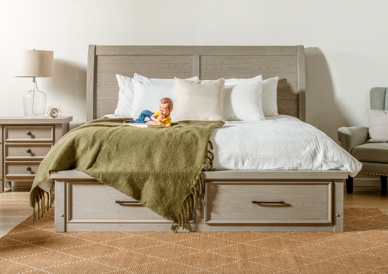 Rustic styled gray finish bedroom set with storage bed and 3 drawer nightstand, sitting on a beige geometric rug with a gray upholstered chair, olive drag green blanket, and glass table lamp.