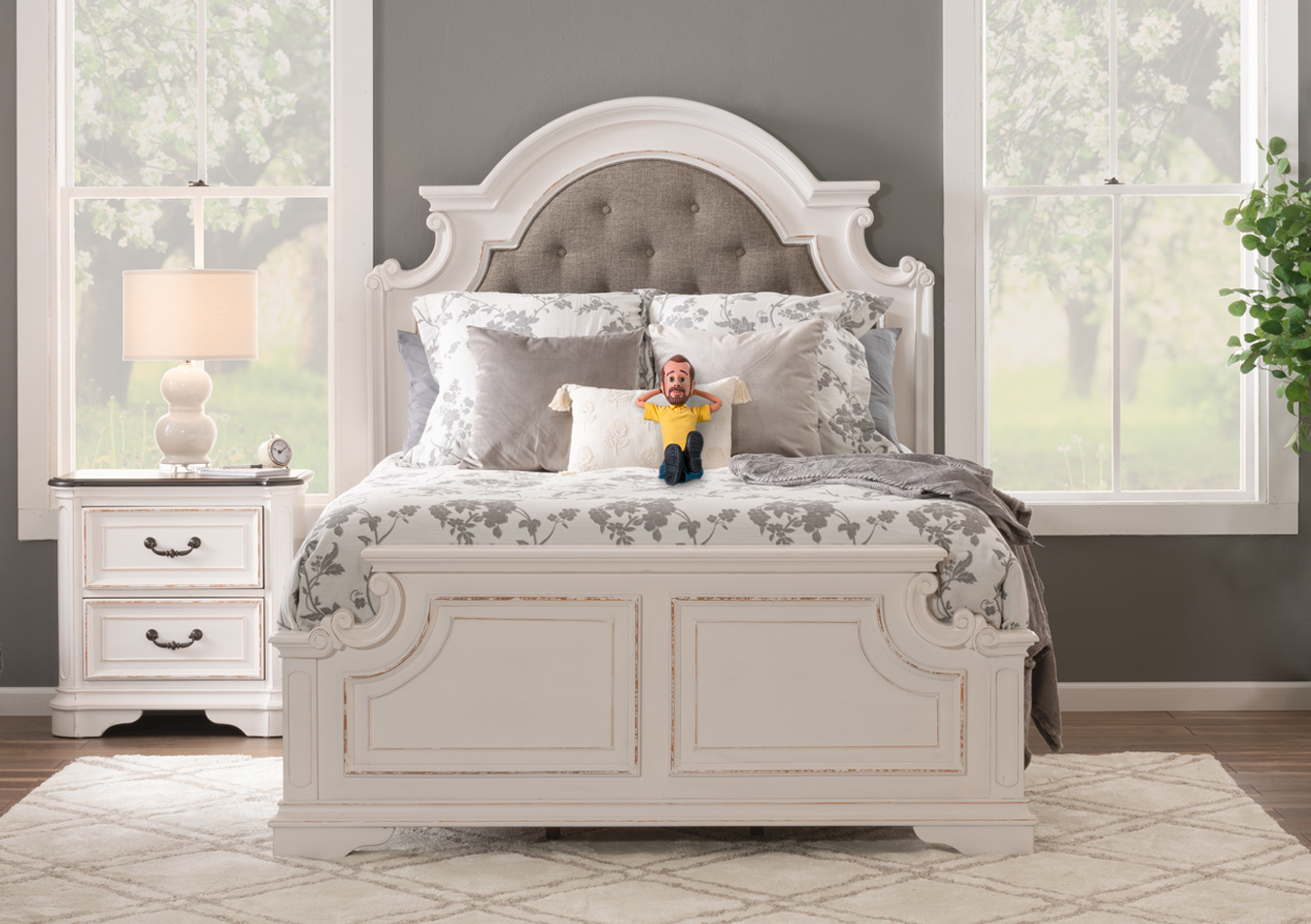 Cottage style distressed white finish with gray accents and brass hardware bedroom set with an upholstered and tufted headboard bed, and 2 drawer nightstand on a white and beige geometric carpet with floral patterned accent pillows and blankets