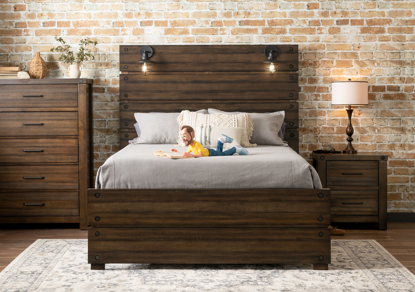Warm brown wooden bedroom set with bed featuring two lamps attached to the headboard, 5 drawer chest, and 2 drawer nightstand, atop a faded beige and gray oriental rug with traditional wooden lamp.