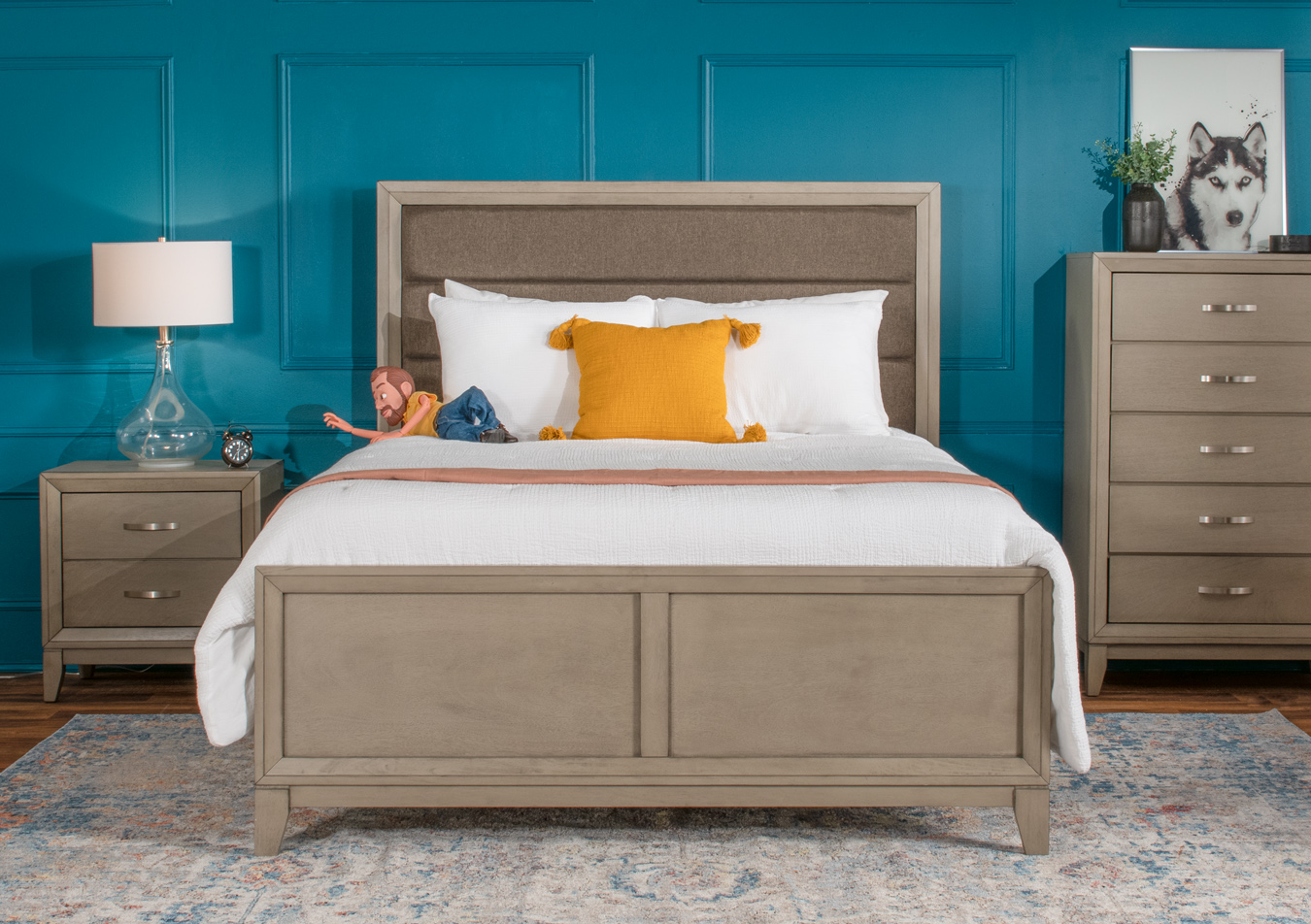 Stunning gray bedroom set with a gray upholstered headboard bed, 5 drawer chest, and 2 drawer nightstand, on an abstract carpet with a decorative glass table lamp and yellow accent pillow.