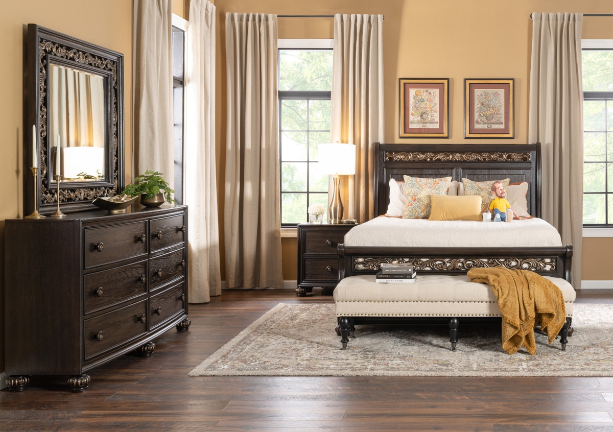 Elegant cherry finish with ornate gold trim bedroom set including a bed, 6 drawer dresser with mirror, and 2 drawer nightstand on top a beige oriental rug with a beige upholstered nailhead bench and golden accent pillows and blankets.