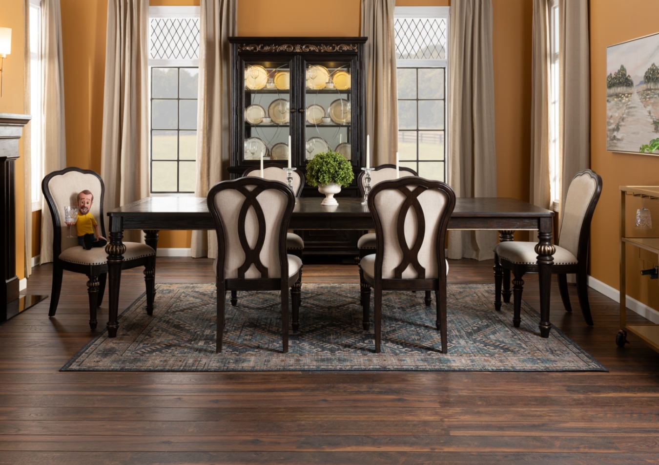 Dining room with brown rectangular dining table with 6 brown and beige full back upholstered dining chairs anchored on a vintage green rug. Brown 2 glass door china cabinet and a gold glass 2 shelf bar cart on wheels.
