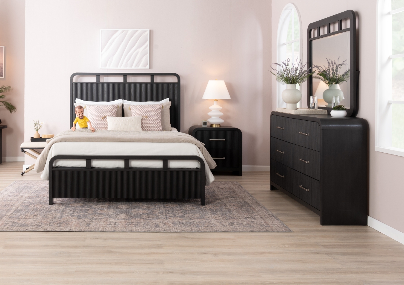 Transitional bedroom set in black featuring a bed with headboard with white sheets and beige comforter, 6 drawer dresser and mirror, and 2 drawer nightstand with white table lamp, all against a white wall and on top a distressed oriental rug.