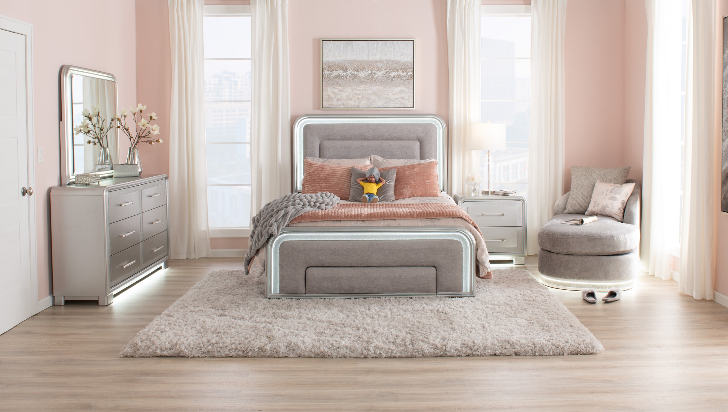 Glam silver bedroom set showcasing a gray upholstered LED backlit bed with headboard, 6 drawer dresser with LED backlit mirror and base, and nightstand with base lighting, all against a light pink room with natural floors and white shag area rug.