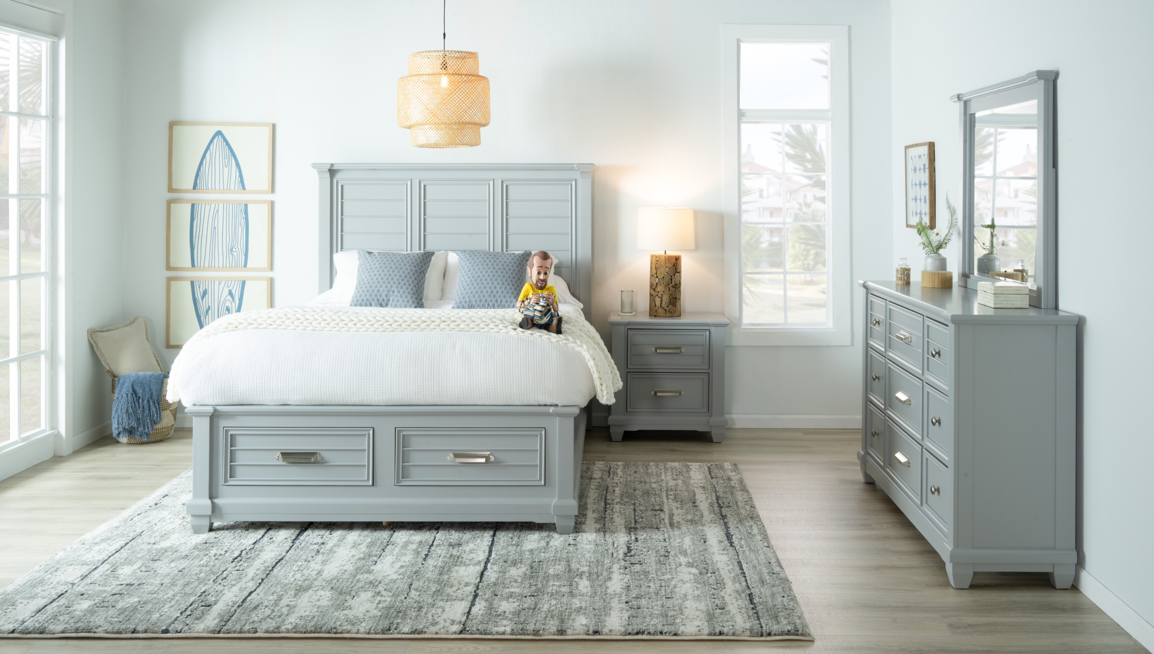 Coastal gray bedroom set with silver hardware featuring a storage bed with panelled headboard, 9 drawer dresser with beveled mirror, and 2 drawer nightstand with wooden lamp on top, all set against a light blue wall and grayish brown flooring.
