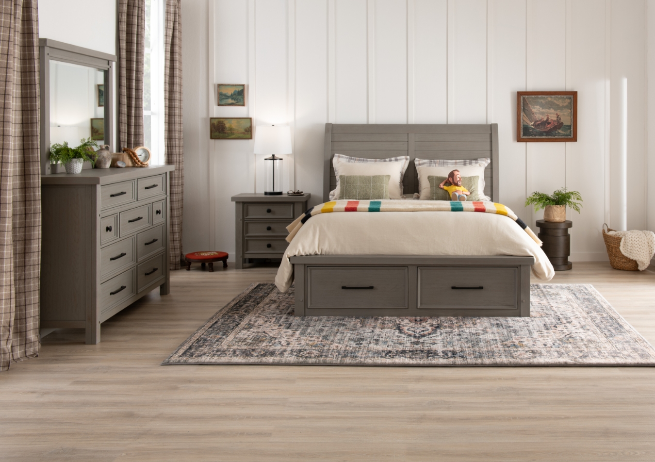 Rustic styled gray finish bedroom set with storage bed and 3 drawer nightstand, sitting on a beige geometric rug with a gray upholstered chair, olive drag green blanket, and glass table lamp.