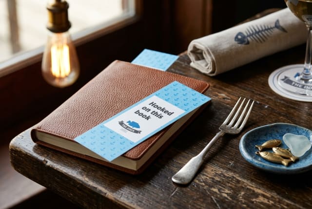 A tall, custom-printed bookmark featuring an oyster tasting guide resting on a wooden table next to a folded dinner napkin.