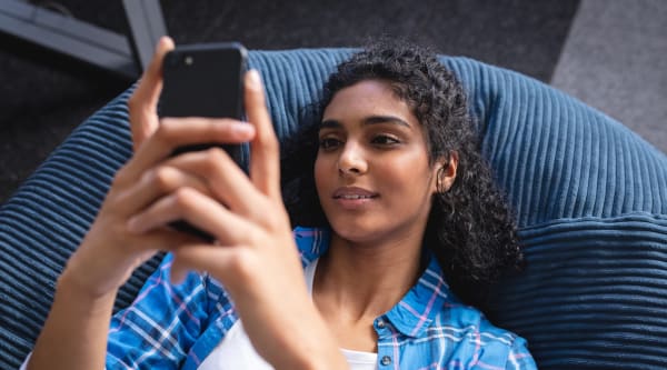 happy mixed race businesswoman chilling in office relaxing space lying in beanbag using smartphone. independent creative design business.