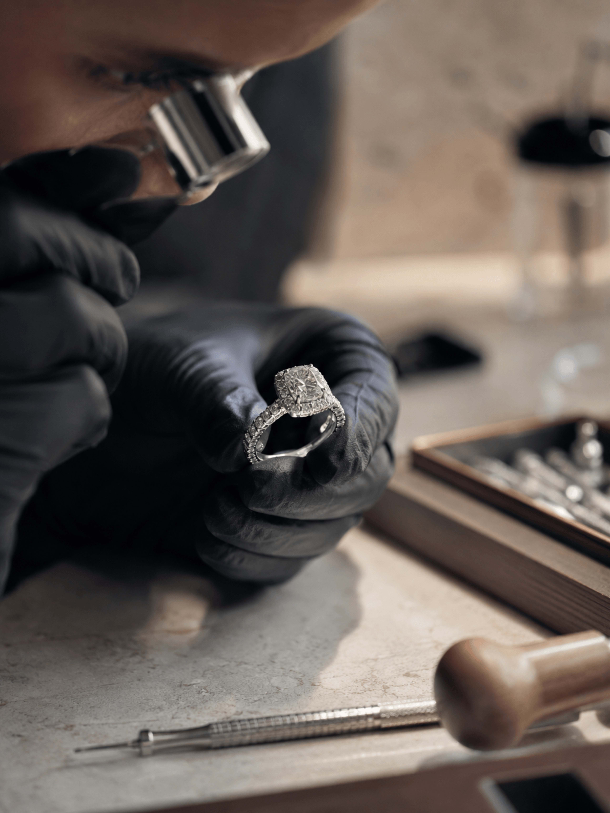 A jeweller examining a halo ring through a loupe at the bench, tools laid out beside them