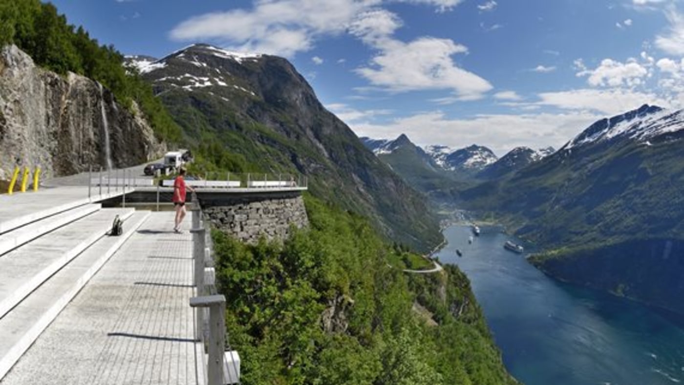 Geiranger. Trollstigen. Mann ser på utsikt. Stupbratt. Turistbåter skimtes på fjorden. Fjell med snø på toppene. 