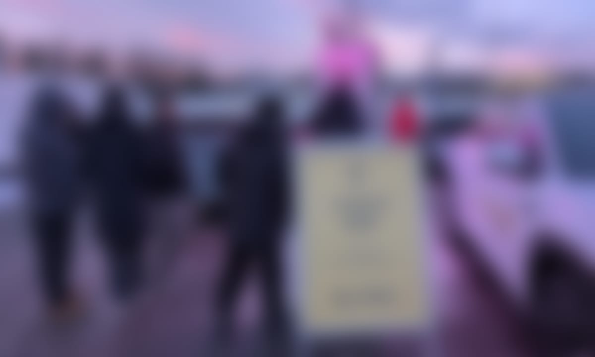 Skilt med "ladetest pågår" foran ladestolpe, hvor et knippe mennesker følger nøye med når e bil lades.