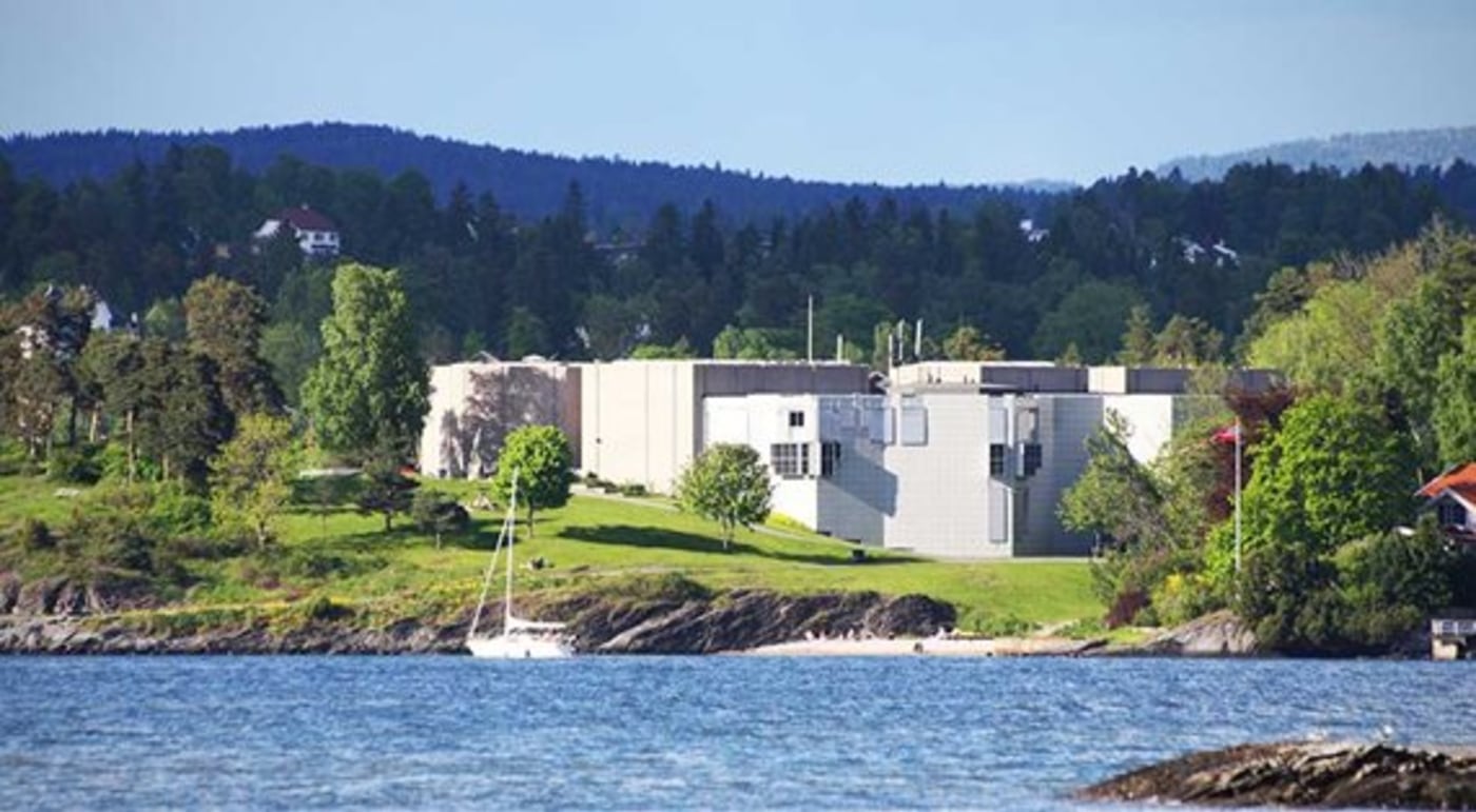 Henie-Onstad Kunstsenter. Stort murbygg ligger i kupert terreng ved vannet. Frodig vegetasjon rundt. 