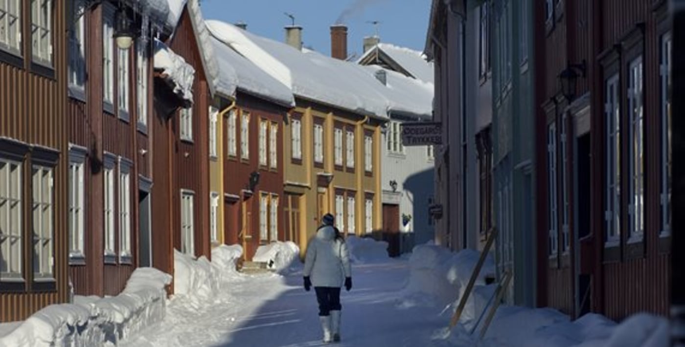 Dame går i idyllisk vinterkledd gate. Tett i tett med trehus på hver side. 